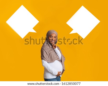 Pregnant afro muslim woman in hijab posing over yellow background with two empty speech bubbles. Woman choosing name, guessing gender of baby