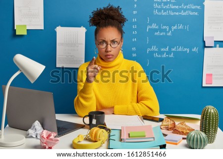 Photo of angry woman with Afro hair points at you, blames in wrong doing, looks seriously at camera, wears casual clothing, poses in coworking space of study room, prepares home assigment or project