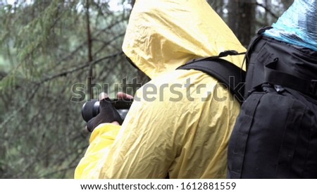 Side view of a youn man standing among trees and taking pictures of summer forest. Stock footage. Hiker in yellow jacket with a camera in his hands in the forest.