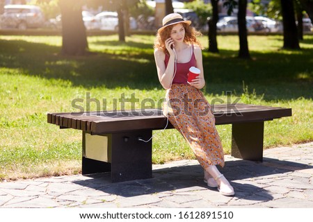 Woman charging her phone on solar panel charger incorporated in to sitting bench, drinking takeaway coffee while talking to someone via her smart phone. Modern technology, alternative energy coconcept Royalty-Free Stock Photo #1612891510