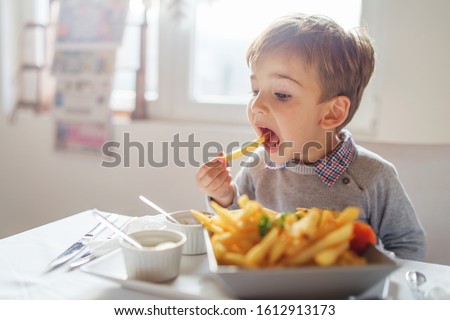 Portrait of small little cute caucasian boy kid eating french fries potato chips at the table in the restaurant or at home three or four years old Royalty-Free Stock Photo #1612913173