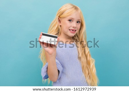 Portrait of little caucasian girl in blue t-shirt with long blonde hair holds a credit card isolated on blue background