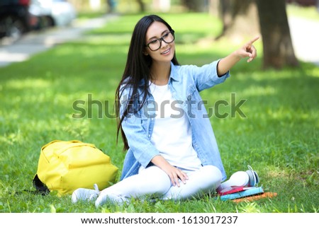 Beautiful young woman with book, headphones and backpack sitting on green grass in the park