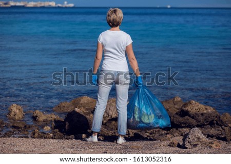 Woman volunteer in white t-shirt with big blue bag collecting garbage on beach. Environmental pollution concept