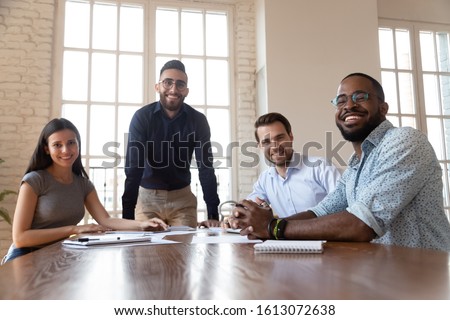 Portrait of happy millennial multiethnic work team sit at office desk brainstorming at meeting looking at camera, successful smiling diverse businesspeople posing in boardroom gather at briefing Royalty-Free Stock Photo #1613072638