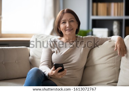 Portrait of happy middle-aged female sit rest on cozy sofa in living room look ta camera using modern cellphone, smiling senior woman smartphone user relax on couch at home posing, technology concept