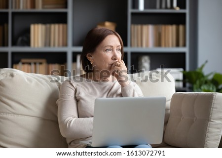 Pensive middle-aged woman sit on couch in living room using laptop look in distance thinking or pondering, thoughtful senior female distracted lost in thoughts feel lonely or sad at home Royalty-Free Stock Photo #1613072791