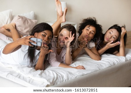 Overjoyed multiracial millennial girls lie on bed have fun pose for self-portrait picture on cell, smiling multiethnic diverse female friends take selfies celebrate hen pajama party at home together