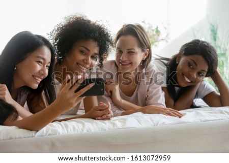 Happy multiracial female friends relax on bed smile pose for self-portrait picture on smartphone, overjoyed multiethnic millennial girls celebrate pajama hen party at home taking selfies on cell