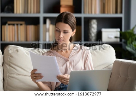 Focused young woman sit on couch in living room working on laptop considering paperwork correspondence, serious millennial female read paper contract or notice use computer at home Royalty-Free Stock Photo #1613072977