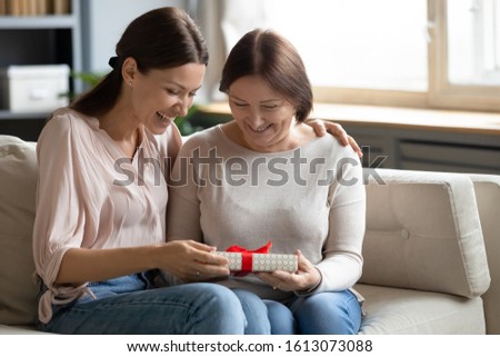 Overjoyed grownup daughter sit on couch with smiling senior mother greeting giving wrapped gift, caring adult girl child congratulate make birthday surprise to middle-aged mom, celebrating at home Royalty-Free Stock Photo #1613073088