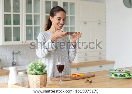 Smiling mixed race woman holding smartphone, making photo of party or romantic dinner preparation process. Happy young female food blogger recording video of salad recipe in modern kitchen at home.