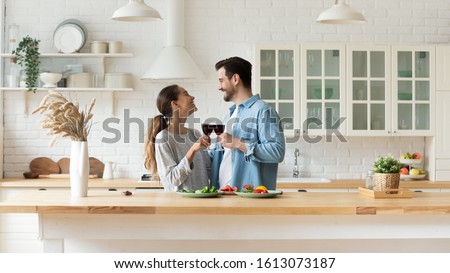 Happy mixed race young woman cheering glasses of red wine with smiling man, enjoying romantic family time in modern kitchen. Excited married couple spending free weekend time together at home. Royalty-Free Stock Photo #1613073187