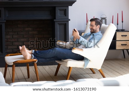 Full length happy young man in eyeglasses relaxing on comfortable armchair, putting legs on ottoman, using smartphone. Smiling millennial guy resting in modern living room, chatting in social network. Royalty-Free Stock Photo #1613073190