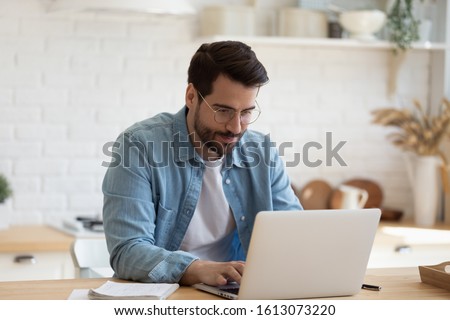 Head shot young businessman sitting at table with computer, working remotely, writing email or reading news. Young man attending distant educational courses, planning job or communicating online. Royalty-Free Stock Photo #1613073220