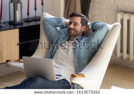 Peaceful young man daydreaming in comfortable armchair with computer on knees. Calm guy taking break after working with laptop, controlling fatigue or stress, resting alone in living room at home. Royalty-Free Stock Photo #1613073262