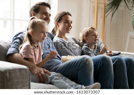 Happy young family with small kids sit relax on couch in living room eating snack popcorn watching video together, smiling parents with little children rest at home on sofa enjoy movie time on TV