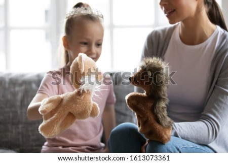 Loving mum or nanny sit on couch engaged in funny childish activity with little girl child at home, caring mother enjoy spending time playing with fluffy hand toys together with small daughter