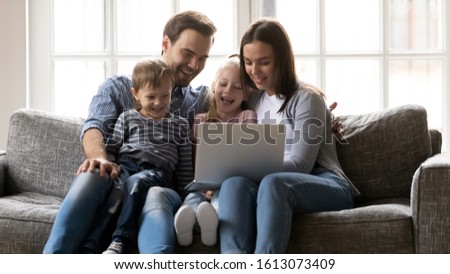 Overjoyed young Caucasian family with little kids relax on sofa watch funny video on laptop together, happy parents with small children rest on couch in living room laugh use computer at home