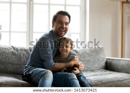 Overjoyed young dad sit on couch play with excited small son enjoy weekend at home, happy father relax on sofa in living room have fun spend time with little boy child together, family bonding concept