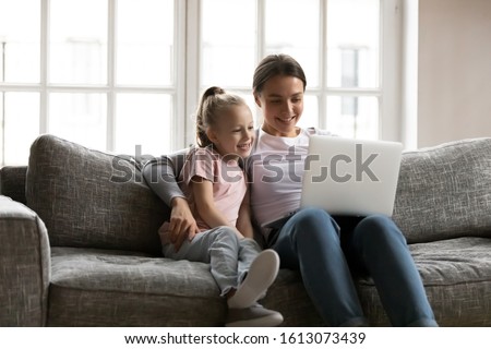 Happy young mum or nanny embrace cute little girl sit relax on couch in living room use laptop together, smiling mother and small daughter rest on sofa at home watch funny video on computer