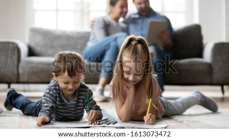 Happy little kids siblings lying on warm floor in living room drawing picture with colorful pencils, smiling small children brother and sister have fun painting in album playing at home together