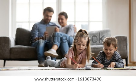 Little kids lying on floor in living room paint picture with colorful pencils, parents relax on couch enjoy weekend together, small children brother and sister drawing at home, family time concept