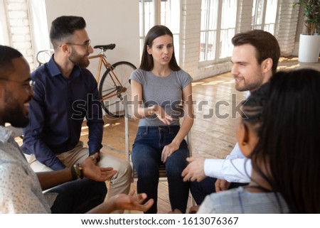 Caucasian millennial woman sit in circle involved in team building activity talk share thoughts with multiethnic participants, young girl speak engaged in group therapy session or treatment in studio Royalty-Free Stock Photo #1613076367