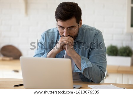 Head shot thoughtful businessman in eyewear looking at computer screen, sitting at table at home. Pensive confused young man thinking of problem solution stuck with task, working with laptop remotely. Royalty-Free Stock Photo #1613076505