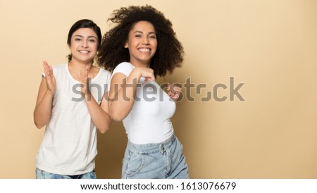 Joyful diverse multicultural female friends dancing, clapping hands, having fun together near blank empty copy space. Happy indian ethnicity and african american women isolated on yellow background.