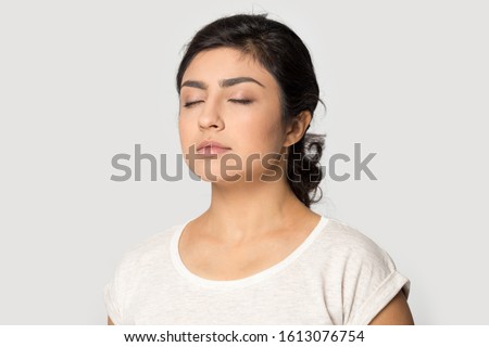 Half side view peaceful millennial indian girl standing with closed eyes, feeling tranquil mindful, meditating, reducing stress, taking break pause, breathing fresh air, isolated on grey background.