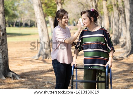 Asian daughter using wet wiping face for her senior mother,caring woman wipe sweat for elderly people by tissue paper while walking with walker in outdoor at park,support care clean sweat on skin face