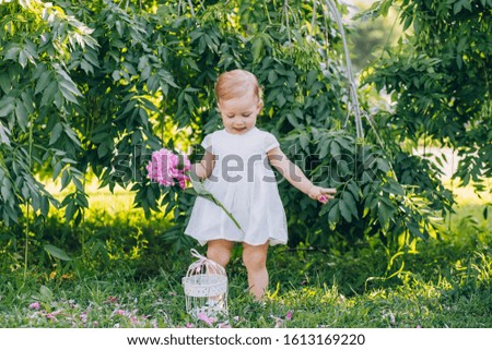 Little cute girl child in a white dress with a pink peony in hands in the park. Portrait of a child outdoors on a sunny summer day. Nice girl holding flowers in her hands.