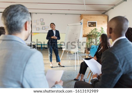 Smiling Asian business trainer standing near whiteboard. Group of workers sitting and listening speaker. Business meeting concept
