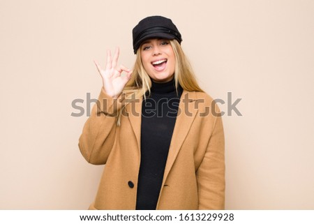 young blonde woman feeling successful and satisfied, smiling with mouth wide open, making okay sign with hand against flat wall