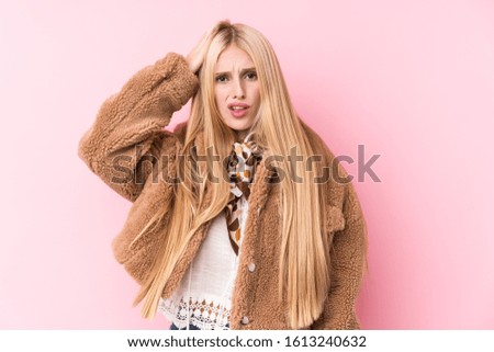 Young blonde woman wearing a coat against a pink background tired and very sleepy keeping hand on head.