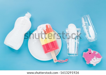 Bottle with washing agent and sponge in the form of ice cream and glass on soapy foam background. Washing dishes concept. Flat lay, Top view.