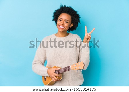 Young african american woman playing ukelele isolated joyful and carefree showing a peace symbol with fingers.