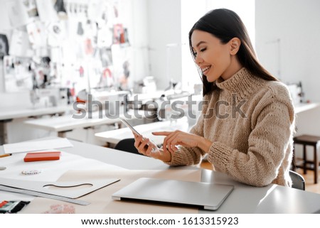 Preparation for work at the workplace. Asian woman seamstress tailor examines the details of her new project on the cell phone. Her own studio interior background. Tailor school. Lovely freelance work