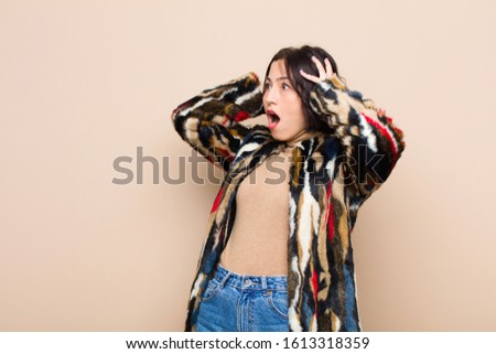 young pretty woman with open mouth, looking horrified and shocked because of a terrible mistake, raising hands to head against beige wall