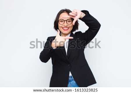 young businesswoman feeling happy, friendly and positive, smiling and making a portrait or photo frame with hands against white wall