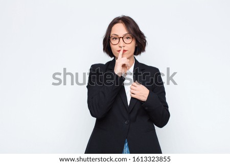 young businesswoman looking serious and cross with finger pressed to lips demanding silence or quiet, keeping a secret against white wall