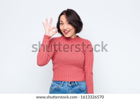 young pretty woman feeling successful and satisfied, smiling with mouth wide open, making okay sign with hand against white wall