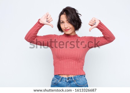 young pretty woman looking sad, disappointed or angry, showing thumbs down in disagreement, feeling frustrated against white wall