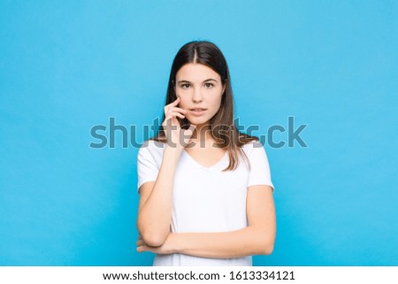 young pretty woman open-mouthed in shock and disbelief, with hand on cheek and arm crossed, feeling stupefied and amazed against blue wall