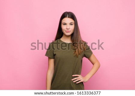 young pretty woman smiling happily with a hand on hip and confident, positive, proud and friendly attitude against pink wall