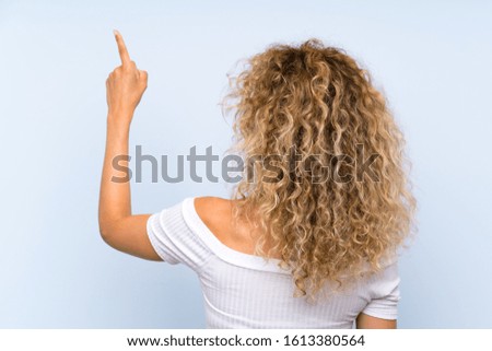 Young blonde woman with curly hair over isolated blue background pointing back with the index finger