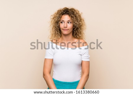 Young blonde woman with curly hair over isolated background making doubts gesture looking side