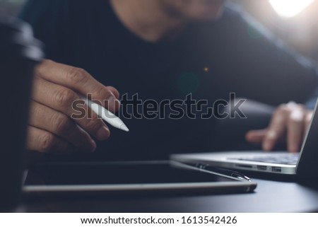Casual business man with stylus pen using digital tablet while working on laptop computer in office. Web designer working his web design project, dark tone, close up. Business and technology concept