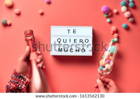 Text "Te quero mucho" means I love you so much in English. Lightboard with text in Latino female hands, flat lay on pink background. Bottle of champagne, flute glass with decorative bubbles.